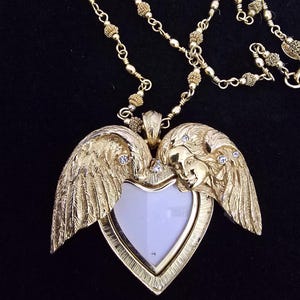 Puede incluir: Collar dorado con un colgante en forma de coraz&oacute;n con alas de &aacute;ngel y una cara. El colgante tiene un centro blanco y est&aacute; adornado con peque&ntilde;os cristales transparentes. La cadena est&aacute; hecha de delicados eslabones dorados.