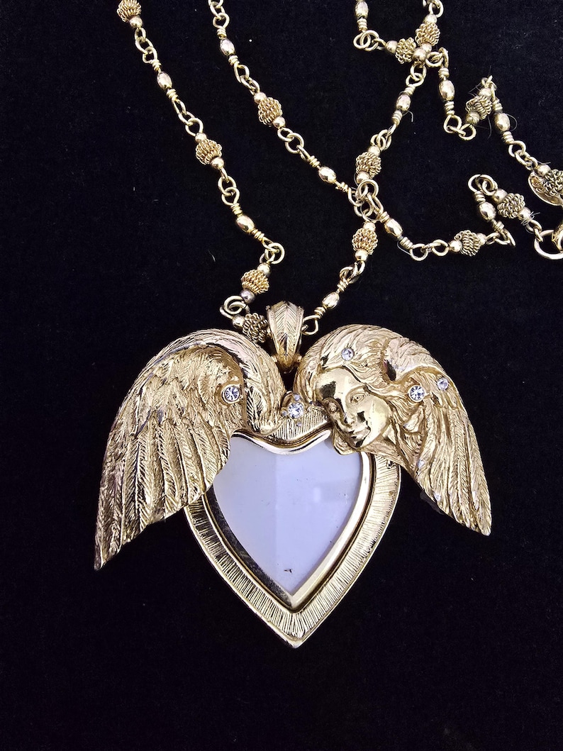 Puede incluir: Collar dorado con un colgante en forma de coraz&oacute;n con alas de &aacute;ngel y una cara. El colgante tiene un centro blanco y est&aacute; adornado con peque&ntilde;os cristales transparentes. La cadena est&aacute; hecha de delicados eslabones dorados.