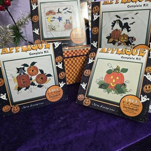May include: Four square Applique Complete Kit packages featuring Halloween themes with pumpkins, ghosts, cats, and bats. Each package includes a graphic of the finished design, the text Applique Complete Kit, and a circular label stating Free E-Z Paint Iron-On Transfer Included.