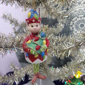 May include: A Christmas ornament featuring a ceramic elf holding a decorated Christmas tree. The elf wears a red outfit with a white collar and a colorful hat. The tree is green with colorful lights and a gold star. The ornament hangs from a gold string.