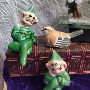 May include: Three ceramic figurines, two green elves with pointy hats and one brown bird, sitting on a wooden box with intricate carvings. The elves have closed eyes and are sitting with their legs crossed. The bird has a brown and white striped pattern.