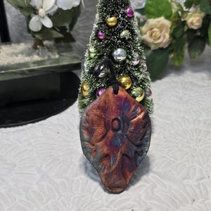 May include: A teardrop-shaped pendant with a textured, metallic finish in shades of copper, bronze, and deep blue. The pendant features a central oval indentation and is suspended by a black cord, showcasing a leaf-like pattern across its surface.