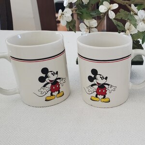 Op de afbeelding: Twee witte keramische mokken met zwarte, rode en witte randen. Elke mok heeft een cartoonafbeelding van Mickey Mouse in zwart, rood, geel en wit. De mokken hebben een gebogen handvat.