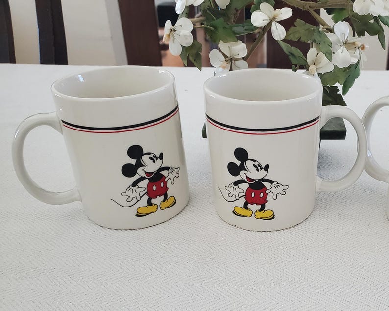 Op de afbeelding: Twee witte keramische mokken met zwarte, rode en witte randen. Elke mok heeft een cartoonafbeelding van Mickey Mouse in zwart, rood, geel en wit. De mokken hebben een gebogen handvat.