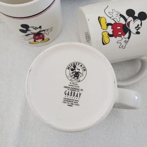 Op de afbeelding: Witte keramische mokken met Mickey Mouse-illustraties. E&eacute;n mok heeft een rood-zwarte streep bij de rand. De mokken tonen Mickey Mouse in verschillende poses, met zijn rode broek en gele schoenen. De bodem van &eacute;&eacute;n mok toont het Mickey & Co. logo.