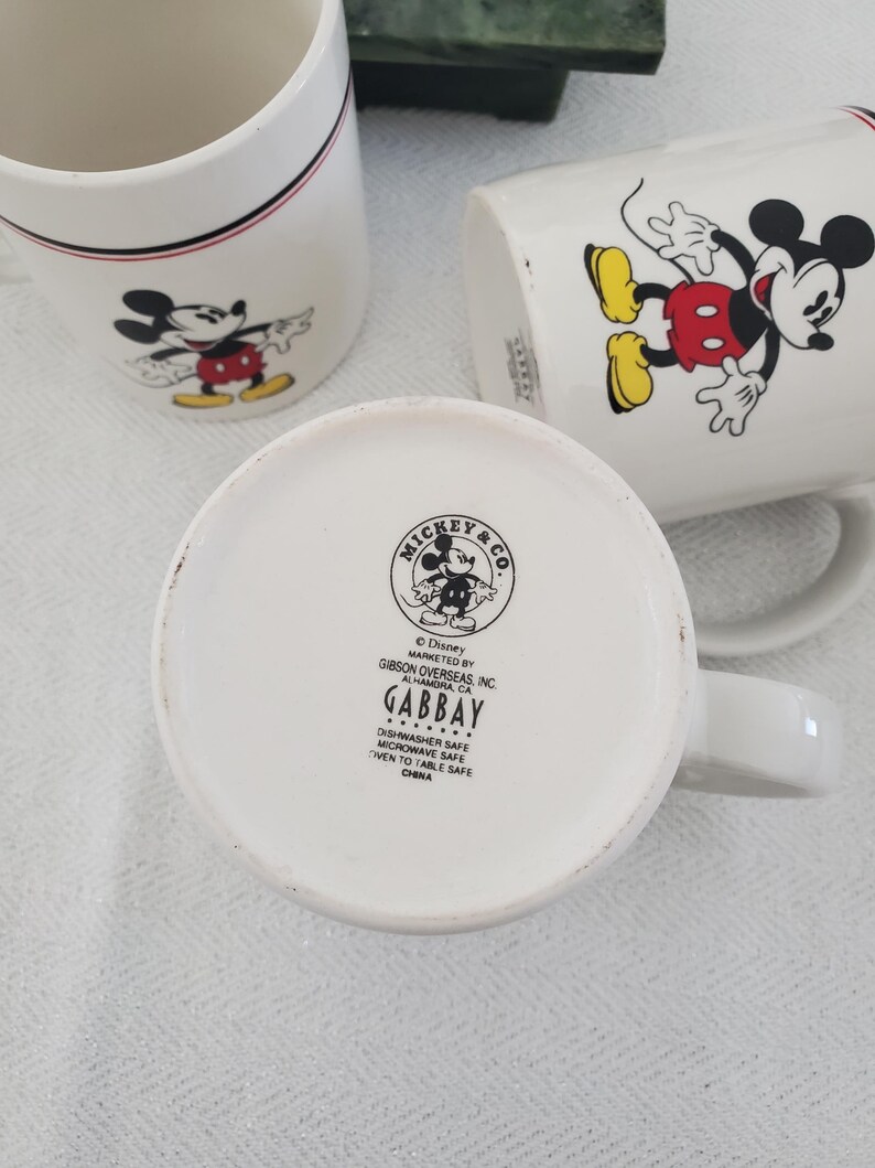 Op de afbeelding: Witte keramische mokken met Mickey Mouse-illustraties. E&eacute;n mok heeft een rood-zwarte streep bij de rand. De mokken tonen Mickey Mouse in verschillende poses, met zijn rode broek en gele schoenen. De bodem van &eacute;&eacute;n mok toont het Mickey & Co. logo.