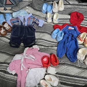 May include: A collection of doll clothing and accessories including a plaid shirt, denim shorts, a blue cap, a red hat, a blue and red jogger outfit with the text Jogger, a pink gingham dress with white lace trim, and various pairs of doll shoes.