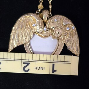 Puede incluir: Collar con colgante dorado con un dise&ntilde;o en forma de coraz&oacute;n, alas de &aacute;ngel y caras. El colgante tiene un centro blanco y mide aproximadamente 3,8 cm de ancho. Peque&ntilde;as piedras transparentes est&aacute;n engastadas en las alas. El collar est&aacute; en una cadena dorada.