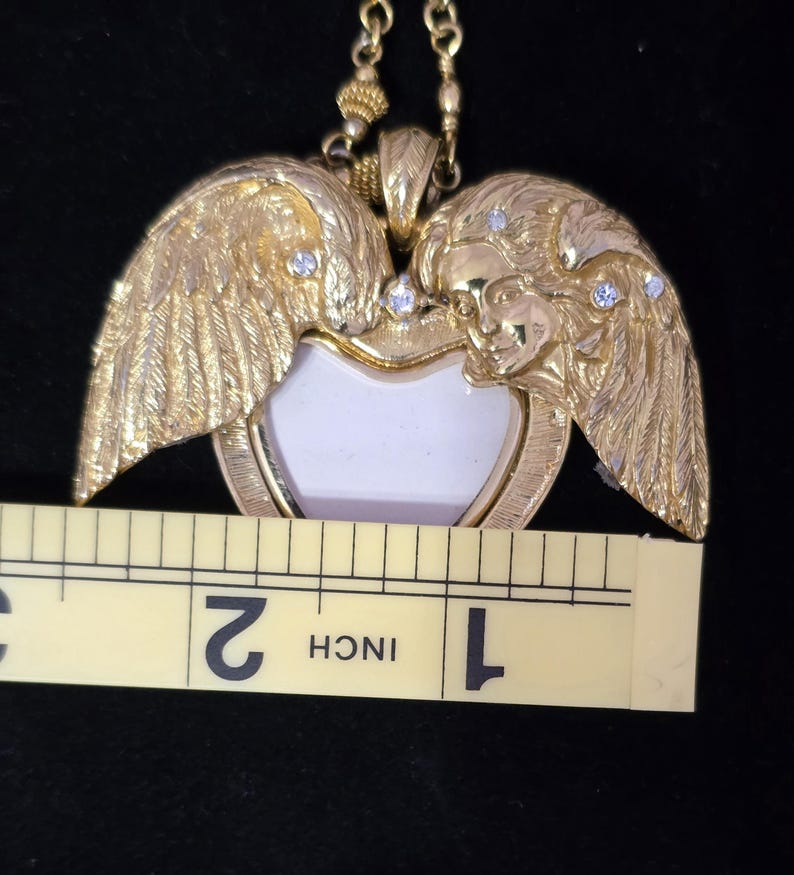 Puede incluir: Collar con colgante dorado con un dise&ntilde;o en forma de coraz&oacute;n, alas de &aacute;ngel y caras. El colgante tiene un centro blanco y mide aproximadamente 3,8 cm de ancho. Peque&ntilde;as piedras transparentes est&aacute;n engastadas en las alas. El collar est&aacute; en una cadena dorada.