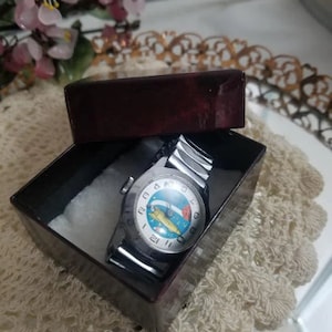 May include: A vintage wristwatch with a silver band and a colorful face featuring a rocket ship design, presented in a dark burgundy box. The watch face displays numbers and a unique illustration. The box is lined with white fabric.
