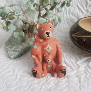 May include: A terracotta-colored ceramic figurine depicting a large bear holding two smaller bear cubs. The bears feature painted details in black, white, and turquoise, including geometric patterns on the chest and paw prints on the feet.