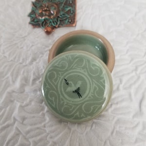 May include: A small, green ceramic trinket box with a lid. The lid has a white bird design with a floral pattern around it.