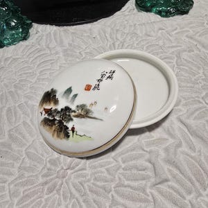 May include: A round white porcelain trinket box with a removable lid. The lid features a hand-painted landscape scene with mountains, trees, and a figure, along with black Chinese calligraphy and a small red square seal. The rim is gold-trimmed.