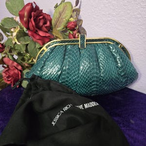 May include: A teal green clutch bag with a textured snakeskin pattern, featuring a gold-tone metal frame and clasp. Accompanied by a black fabric dust bag with the white printed text JESSICA RICH STEVE MADDEN visible on the front surface.