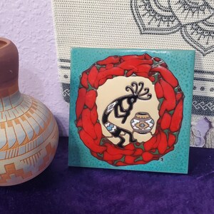 May include: Square ceramic tile with a turquoise background, featuring a Kokopelli figure and a small pot. The Kokopelli is black with a blue and white design, surrounded by a ring of red chili peppers. Decorative art.