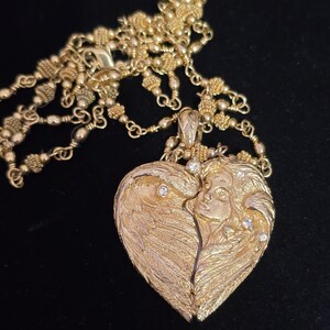 Puede incluir: Un collar dorado con un colgante en forma de coraz&oacute;n. El colgante presenta un dise&ntilde;o detallado de alas de &aacute;ngel y un rostro, adornado con peque&ntilde;as piedras brillantes. La cadena tambi&eacute;n es dorada y tiene un dise&ntilde;o elaborado.