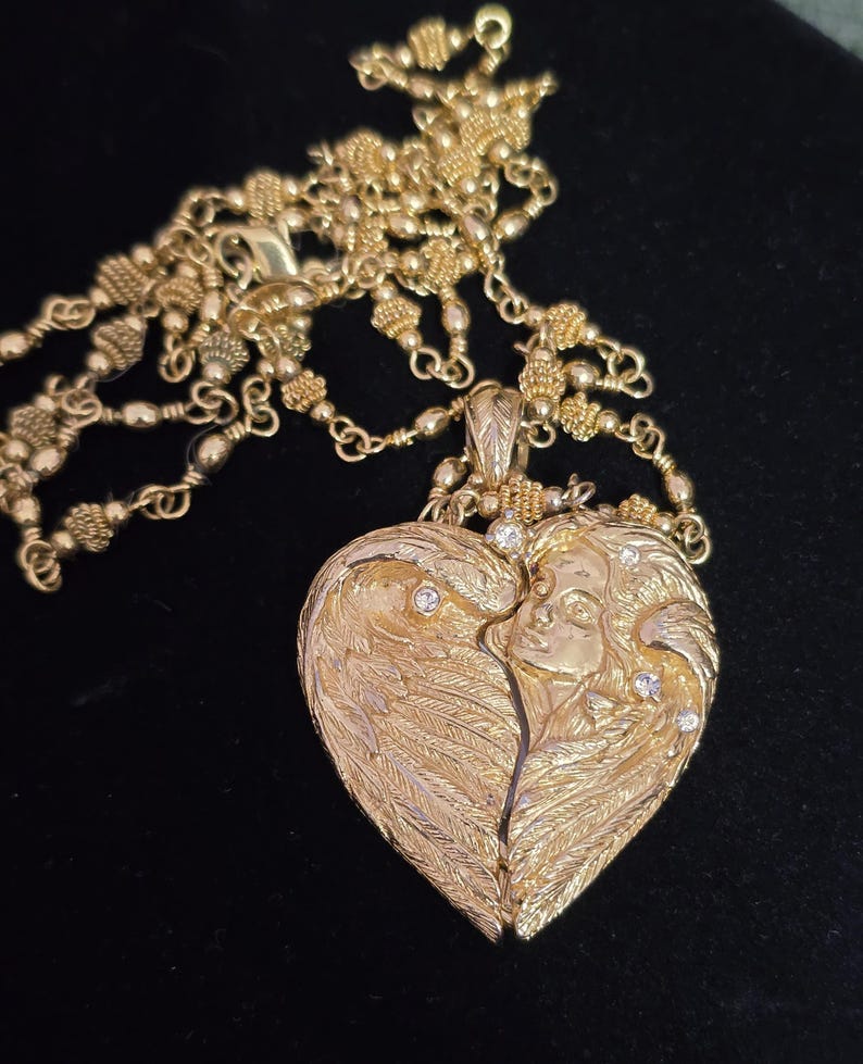 Puede incluir: Un collar dorado con un colgante en forma de coraz&oacute;n. El colgante presenta un dise&ntilde;o detallado de alas de &aacute;ngel y un rostro, adornado con peque&ntilde;as piedras brillantes. La cadena tambi&eacute;n es dorada y tiene un dise&ntilde;o elaborado.