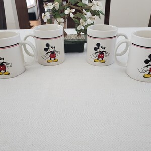 Op de afbeelding: Vier witte keramische mokken met een zwarte, rode en witte rand. Elke mok heeft een kleurrijke illustratie van Mickey Mouse. De mokken zijn gerangschikt op een wit tafelkleed, met een kleine plant op de achtergrond.