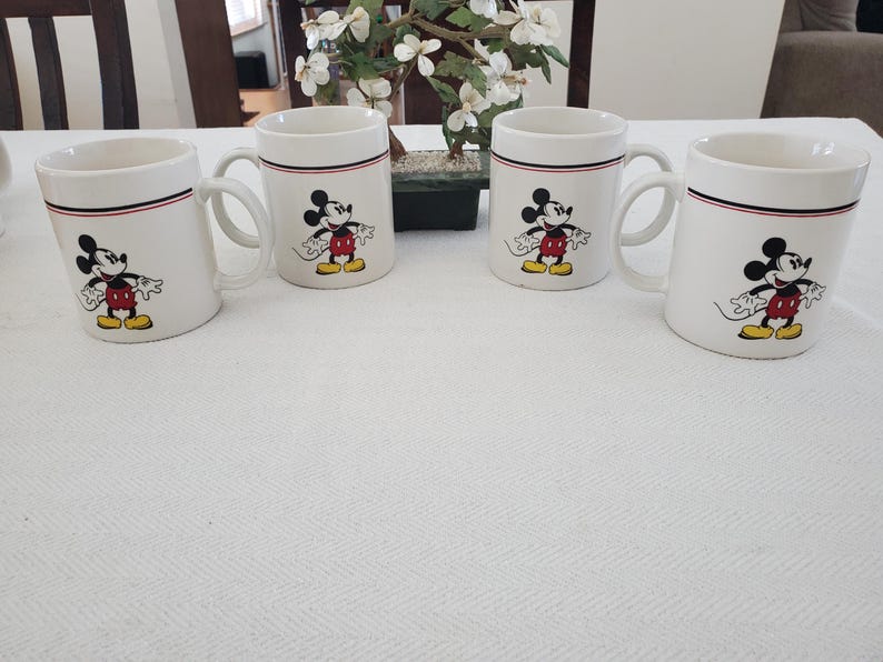 Op de afbeelding: Vier witte keramische mokken met een zwarte, rode en witte rand. Elke mok heeft een kleurrijke illustratie van Mickey Mouse. De mokken zijn gerangschikt op een wit tafelkleed, met een kleine plant op de achtergrond.