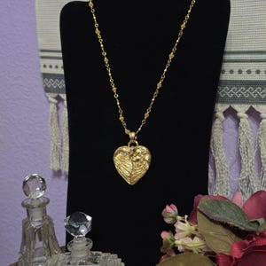 Puede incluir: Un collar dorado con un colgante en forma de coraz&oacute;n, exhibido en un soporte negro. El colgante presenta un dise&ntilde;o detallado. El collar est&aacute; acompa&ntilde;ado de dos frascos de perfume de vidrio y un ramo de flores.