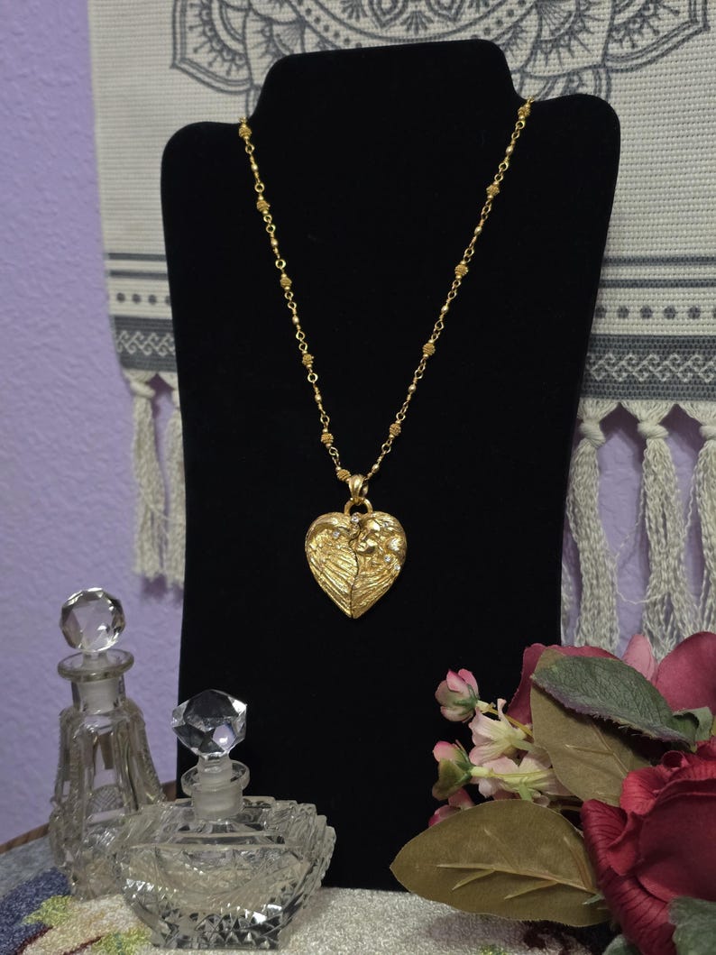 Puede incluir: Un collar dorado con un colgante en forma de coraz&oacute;n, exhibido en un soporte negro. El colgante presenta un dise&ntilde;o detallado. El collar est&aacute; acompa&ntilde;ado de dos frascos de perfume de vidrio y un ramo de flores.