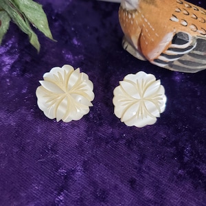 May include: Two white, flower-shaped earrings with carved petal details are displayed on a purple velvet surface. A decorative wooden bird with brown and white markings sits in the background. The earrings are a delicate accessory.