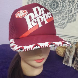May include: A burgundy trucker hat with the Dr Pepper logo in white and red. The brim is decorated with a beaded pattern in white, red, and black. The hat is displayed on a mannequin head.