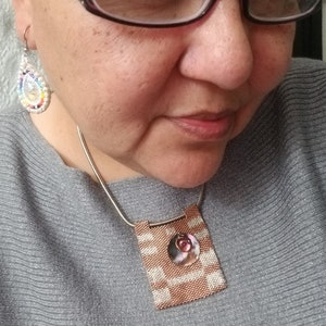 K&ouml;nnte beinhalten: Eine Frau mit Brille und einem grauen Pullover mit einer Perlenkette mit einem quadratischen Anh&auml;nger in Rosa und Wei&szlig;.