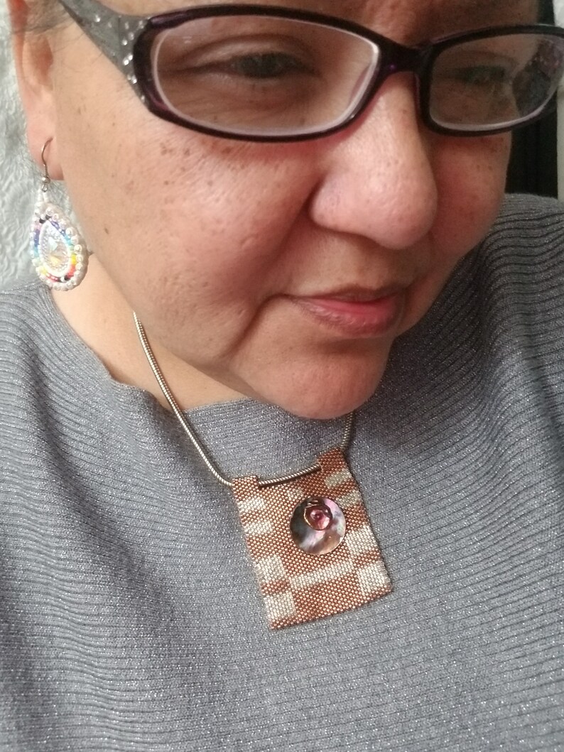 K&ouml;nnte beinhalten: Eine Frau mit Brille und einem grauen Pullover mit einer Perlenkette mit einem quadratischen Anh&auml;nger in Rosa und Wei&szlig;.