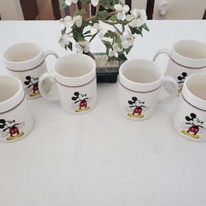 Op de afbeelding: Zes witte keramische mokken met een zwarte en rode rand, elk met een Mickey Mouse-afbeelding. De mokken hebben een handvat en zijn gerangschikt op een wit oppervlak. Een kleine decoratieve plant staat op de achtergrond.