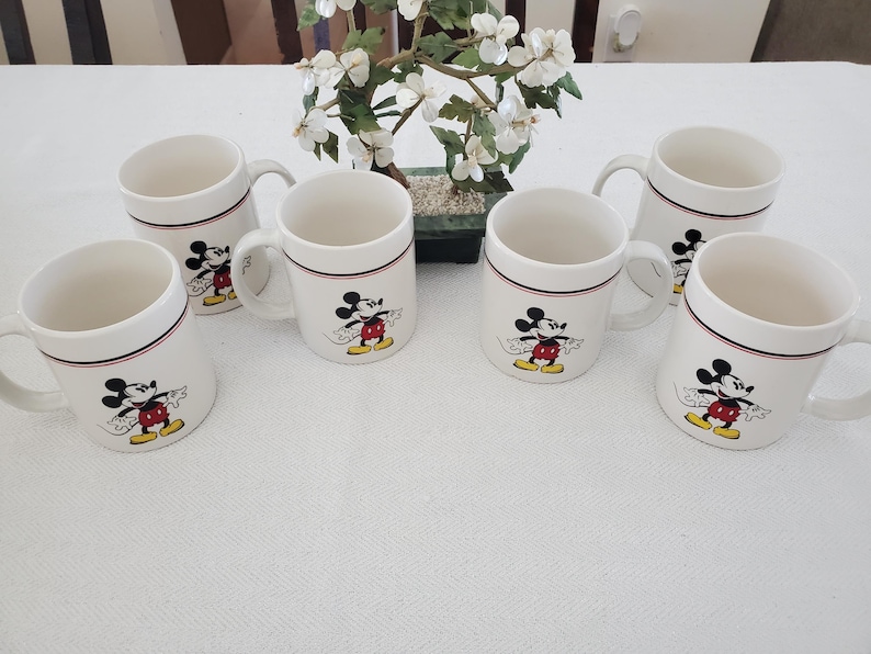 Op de afbeelding: Zes witte keramische mokken met een zwarte en rode rand, elk met een Mickey Mouse-afbeelding. De mokken hebben een handvat en zijn gerangschikt op een wit oppervlak. Een kleine decoratieve plant staat op de achtergrond.