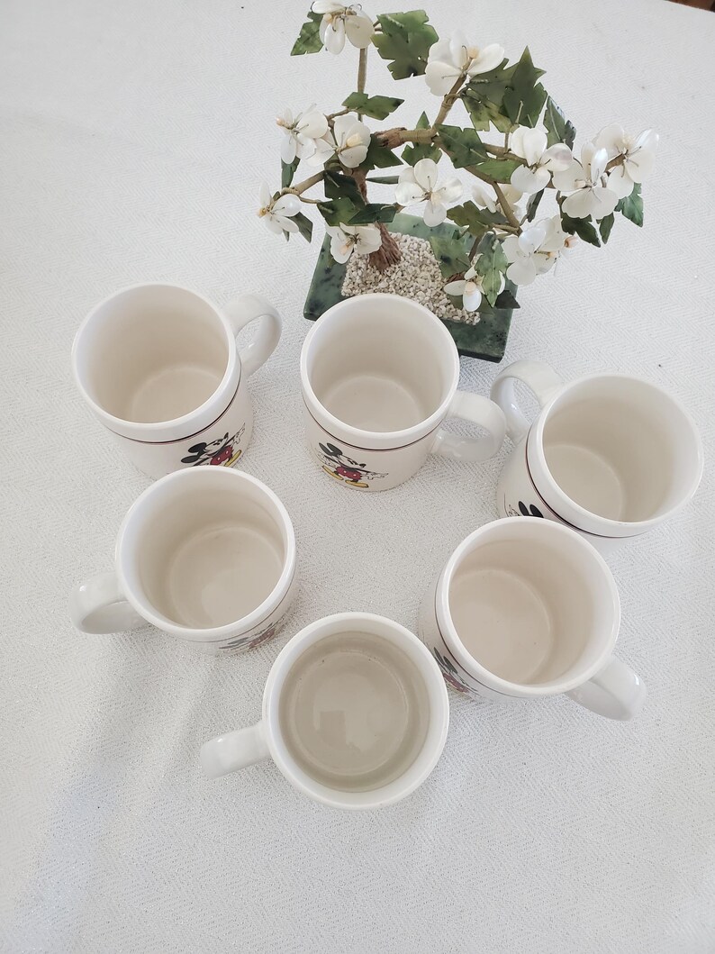 Op de afbeelding: Zes witte keramische mokken met handvatten, elk met een Mickey Mouse-ontwerp. De mokken zijn in een cirkelvormig patroon gerangschikt, waarbij hun binnen- en buitendetails worden getoond. Een decoratief bloemstuk staat op de achtergrond.