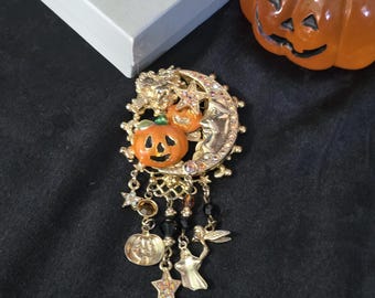 May include: A gold-toned Halloween brooch featuring a crescent moon, pumpkins, and dangling charms. The brooch has an orange pumpkin at the center, with dangling charms including stars, a coin, and a fairy. The brooch is set against a black background.