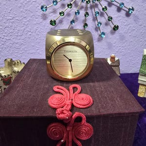 May include: A gold-toned Tiffany & Co. cube-shaped desk clock with a brushed metallic face, black hands, and a smooth finish. The clock sits atop a dark purple fabric-covered box featuring two decorative red knotted fabric closures on the front.