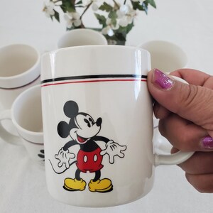 Op de afbeelding: Witte keramische mok met een Mickey Mouse-ontwerp. De mok heeft een zwarte en rode streep bij de rand. Mickey Mouse wordt afgebeeld met een rode broek, gele schoenen en witte handschoenen. Een set mokken is zichtbaar.