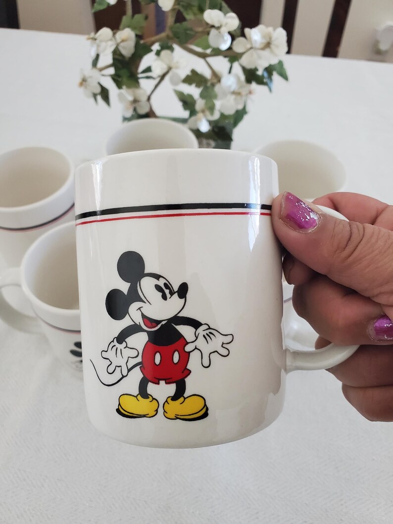 Op de afbeelding: Witte keramische mok met een Mickey Mouse-ontwerp. De mok heeft een zwarte en rode streep bij de rand. Mickey Mouse wordt afgebeeld met een rode broek, gele schoenen en witte handschoenen. Een set mokken is zichtbaar.