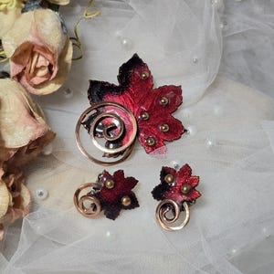 May include: A jewelry set consisting of a brooch and two matching earrings, each shaped like a maple leaf with a gradient of deep red to black, accented with small gold-toned spheres and a prominent copper-colored spiral wire element.
