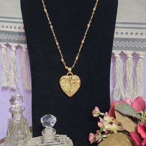 Puede incluir: Un collar dorado con un colgante en forma de coraz&oacute;n con un dise&ntilde;o de &aacute;ngel. El collar se exhibe sobre un soporte negro, con dos frascos de perfume de vidrio decorativos y flores en primer plano.