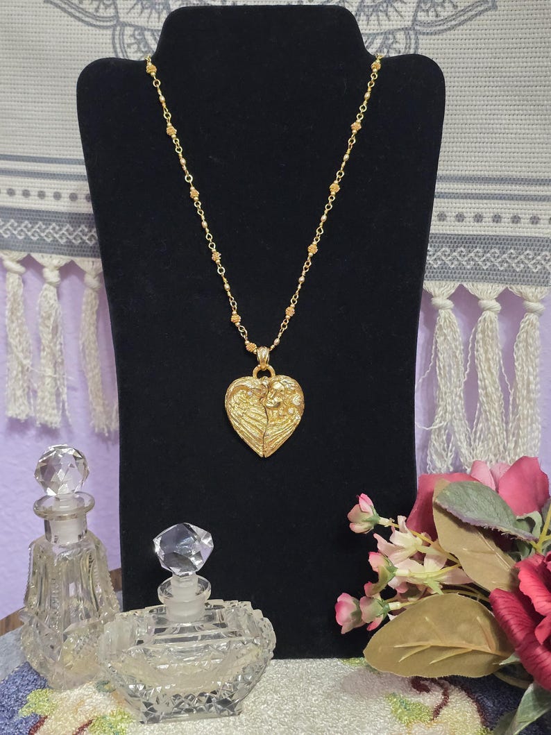 Puede incluir: Un collar dorado con un colgante en forma de coraz&oacute;n con un dise&ntilde;o de &aacute;ngel. El collar se exhibe sobre un soporte negro, con dos frascos de perfume de vidrio decorativos y flores en primer plano.
