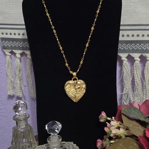 Puede incluir: Un collar dorado con un colgante en forma de coraz&oacute;n. El colgante presenta un dise&ntilde;o detallado de dos figuras y alas. El collar se exhibe en un expositor de joyas negro, con dos frascos de perfume de cristal en primer plano.