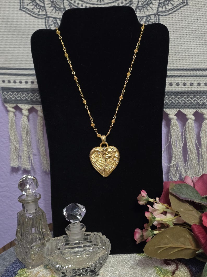 Puede incluir: Un collar dorado con un colgante en forma de coraz&oacute;n. El colgante presenta un dise&ntilde;o detallado de dos figuras y alas. El collar se exhibe en un expositor de joyas negro, con dos frascos de perfume de cristal en primer plano.