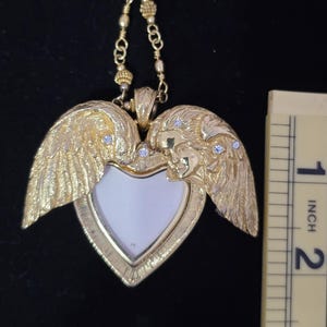 Puede incluir: Un collar con colgante en forma de coraz&oacute;n dorado con alas de &aacute;ngel y una peque&ntilde;a cara a cada lado. El colgante tiene un centro blanco y mide aproximadamente 5 cm de altura. Peque&ntilde;as piedras transparentes est&aacute;n engastadas en las alas.