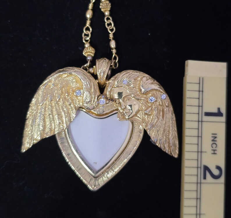 Puede incluir: Un collar con colgante en forma de coraz&oacute;n dorado con alas de &aacute;ngel y una peque&ntilde;a cara a cada lado. El colgante tiene un centro blanco y mide aproximadamente 5 cm de altura. Peque&ntilde;as piedras transparentes est&aacute;n engastadas en las alas.