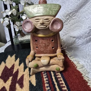May include: A terracotta-colored ceramic figurine of a seated figure with large circular ear ornaments. The figure wears a green headpiece with etched geometric symbols and a brown chest plate featuring white linear patterns and four circular holes.