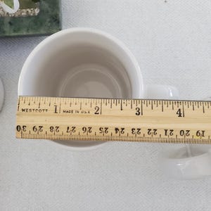 Op de afbeelding: Een witte keramische mok met een oor, gemeten met een houten liniaal. De liniaal toont metingen in inches, met een diameter van de mok van ongeveer 10 cm. De mok staat op een wit oppervlak.