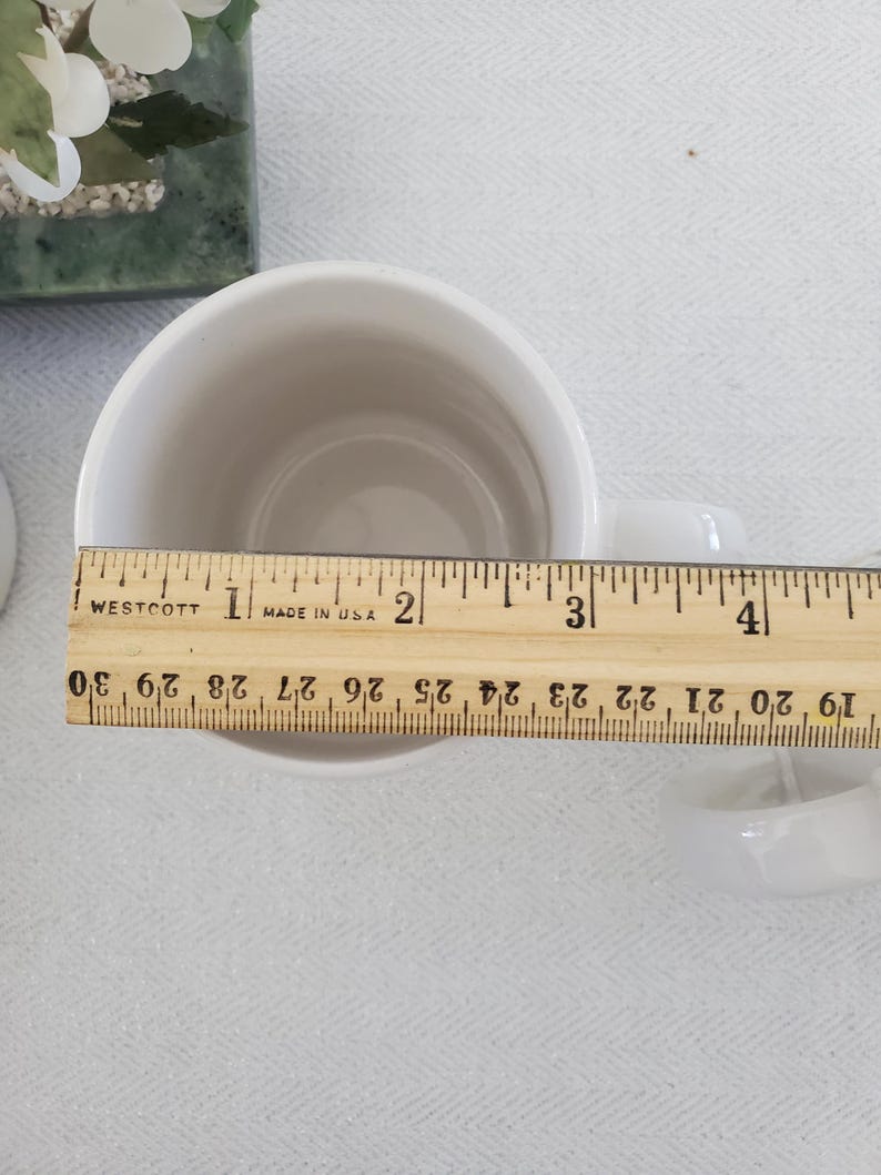 Op de afbeelding: Een witte keramische mok met een oor, gemeten met een houten liniaal. De liniaal toont metingen in inches, met een diameter van de mok van ongeveer 10 cm. De mok staat op een wit oppervlak.