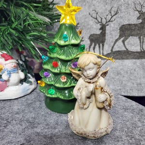 May include: A ceramic Christmas tree with colorful lights and a yellow star topper, next to a small angel ornament holding a candle. The angel is wearing a cream-colored dress with gold accents. The background features a gray fabric with deer silhouettes.
