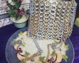 May include: A vintage chainmail purse with silver and gold-tone metal rings. The purse has a chain strap and is displayed on a floral embroidered doily. A small potted plant and decorative earrings are also visible.
