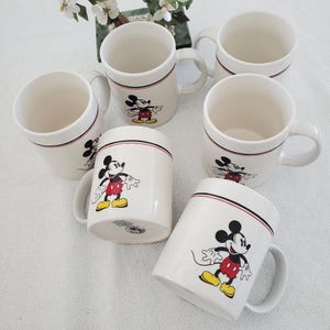 Op de afbeelding: Zes witte keramische mokken met Mickey Mouse. Elke mok heeft een zwarte en rode streep bij de rand en een full-color afbeelding van Mickey Mouse. De mokken hebben een gebogen handvat en staan op een wit oppervlak.