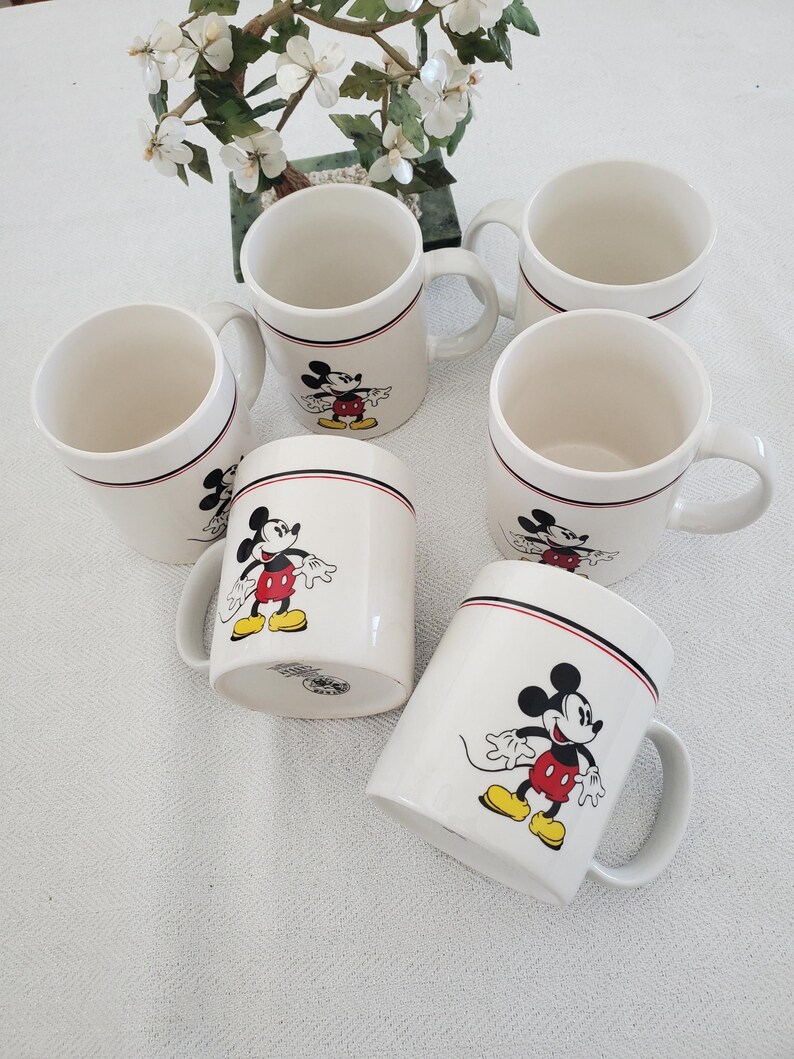 Op de afbeelding: Zes witte keramische mokken met Mickey Mouse. Elke mok heeft een zwarte en rode streep bij de rand en een full-color afbeelding van Mickey Mouse. De mokken hebben een gebogen handvat en staan op een wit oppervlak.